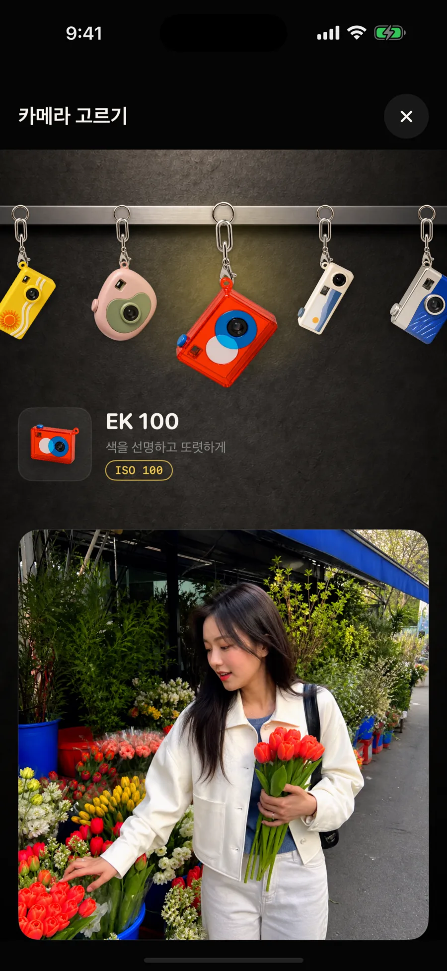Mayica의 카메라 고르기 화면. 벽에 걸린 카메라 키링들과 선택된 EK 100.